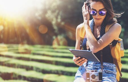 Hipster young girl with bright backpack and vintage camera using tablet or holding gadget, planning travel plan. View tourist traveler with sunglasses on background Parc Laberint Barcelona. Mock up for text messageの写真素材