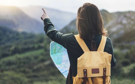 Hipster young girl with bright backpack enjoying sunset on peak of foggy mountain, looking a map and poining hand. Tourist traveler on background valley landscape panoramic view mockup, sunlight in trip in northern spain basque country, mock up for text.の写真素材