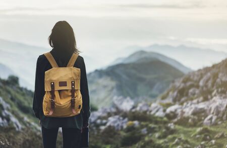 Hipster young girl with bright backpack enjoying sunset on peak of foggy mountain. Tourist traveler on background valley landscape panoramic view mockup. Hiker looking sunlight in trip in northern spain basque country, mock up for text. Girl taking photo of amazing landscape sunset on vintage camera on peak of foggy mountainの写真素材