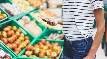 Young woman shopping healthy food in supermarket blur background. Female hands buy products tomato using smartphone in store. Hipster at grocery using smartphone. Person comparing price of produceの写真素材