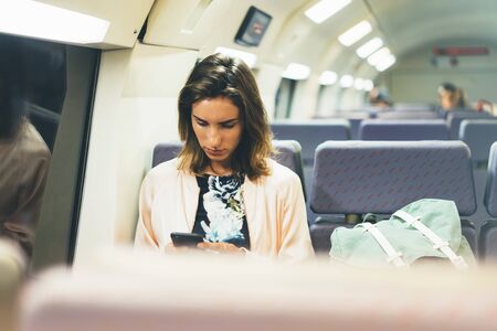 Enjoying travel. Young pretty woman traveling by train sitting near the window using smartphone and looking map. Tourist texting online message and plan route of railway, railroad transport conceptの写真素材