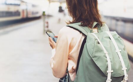 Enjoying travel. Young pretty woman waiting on the station platform with backpack on background electric train using smartphone. Tourist texting message and plan route of railway, railroad transport conceptの写真素材
