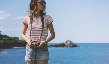 Tourist traveler photographer making pictures seascape on vintage photo camera on background yacht and boat piar, hipster girl enjoying peak mountain and nature holiday, mockup ocean waves view, blurred backdropの写真素材