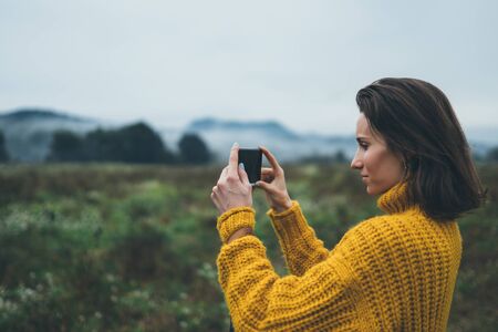 photographer girl hold in hands mobile phone taking photo on smartphone autumn froggy mountain, tourist shooting on photo camera on background landscape, internet online conceptの写真素材