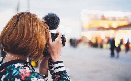 hipster tourist girl hold in hands modern photo camera, take photography click on defocus background bokeh light in evening street attraction, photographer look on camera technology, night city illuminationの写真素材