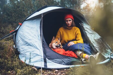 tourist traveler hugging red shiba inu in camp tent on background froggy rain forest, hiker woman with puppy dog in mist nature trip, friendship love concept, girl resting dog together in campsite parkの写真素材