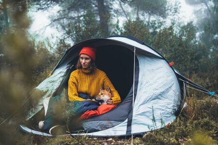 tourist traveler hugging red shiba inu in camp tent on background froggy rain forest, hiker woman with puppy dog in mist nature trip, friendship love conceptの写真素材