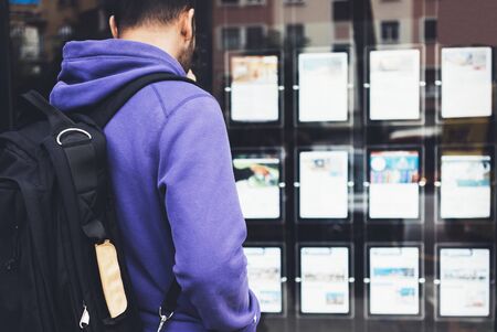 Hipster young man with backpack and map looking ad promo poster hotel choose apartments. Tourist traveler planning route on background advert hostel. Person hiker select hotel in city streetの写真素材