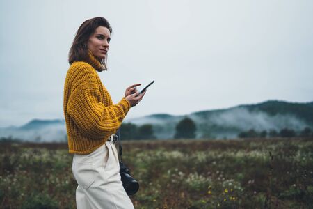 photographer girl hold in hands mobile typing message on smartphone on background landscape autumn froggy mountain, tourist shoot on photo camera using internet online gadget, hobby conceptの写真素材