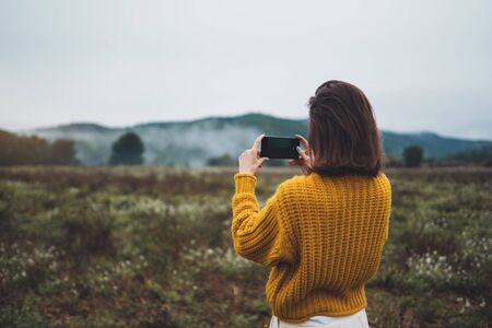 photographer girl hold in hands mobile phone taking photo on smartphone autumn froggy mountain, tourist shooting on photo camera on background landscapeの写真素材