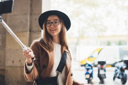 Tourist girl taking selfie photo on smartphone. Girl hipster travels in Barcelona. Holiday concept in Spain street. Traveler in glasses and hat self phone internet technology in europe trip. Copy space mockupの写真素材