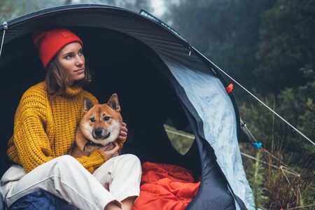 tourist traveler in camp tent hugging red shiba inu on background froggy rain forest, hiker woman with puppy dog in mist nature trip, friendship love concept, relax girl resting dog togetherの写真素材
