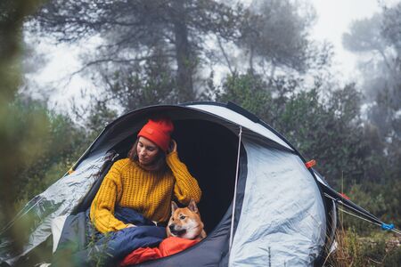 tourist traveler hugging red shiba inu in camp tent on background froggy rain forest, hiker woman with puppy dog in mist nature trip, friendship love conceptの写真素材