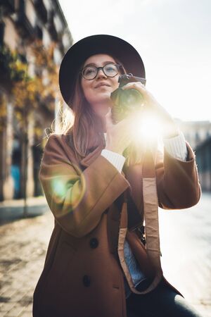 Photographer in glasses with retro photo camera. Tourist portrait girl in hat travels in Barcelona holiday. Sunlight flare street in europe city. Traveler hipster shooting architecture, copy space mockupの写真素材