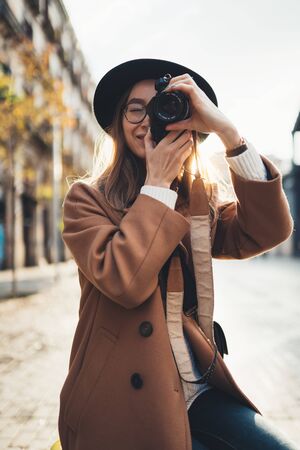 Photographer in glasses take photo on retro camera. Tourist portrait. Girl in hat travels in Barcelona holiday. Sunlight flare street in europe city. Traveler hipster shooting architectureの写真素材