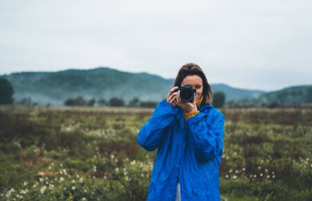 photographer tourist girl in blue raincoat hold in female hands photo camera take photography froggy mountain, traveler shooting autumn nature, click on camera technology, journey landscape vacation concept free spaceの写真素材