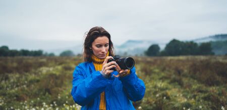photographer tourist girl in blue raincoat hold in female hands photo camera take photography froggy mountain, traveler shooting autumn nature, click on camera technology, journey landscape vacation concept free space backgroundの写真素材