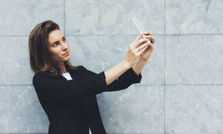 Portrait young businesswomen in black suit using smartphone isolated on background concrete gray wall mockup, pretty hipster manager holding mobile gadget,  girl making selfie photo, connectの写真素材