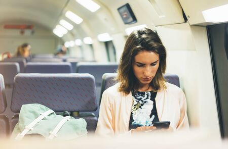 Enjoying travel. Young pretty woman traveling by train sitting near the window using smartphone and looking map in nigth. Tourist texting online message and plan route of railway, railroad transport conceptの写真素材
