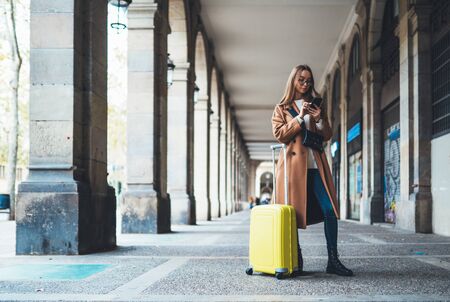 Tourist with suitcase holding smart mobile phone. Girl travels in Barcelona. Holiday concept in Spain street. Traveler hipster in glasses using internet technology in europe trip. Copy space mockupの写真素材