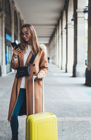 Tourist with suitcase holding smart mobile phone. Girl travels in Barcelona. Holiday concept in Spain street. Traveler hipster in glasses using cellphone internet technology in europe trip. mockupの写真素材