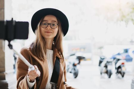 Tourist girl blogger taking photo selfie on smartphone mobile. Girl hipster travels in Barcelona. Holiday concept. Traveler in glasses and hat self cellphone technology in europe trip. Copy space mockupの写真素材