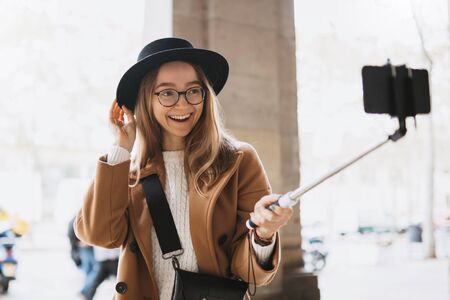 Smiling tourist girl taking photo selfie on smartphone mobile. Blogger hipster travels in Barcelona. Holiday concept in street. Traveler in glasses and hat self cellphone technology in europe tripの写真素材
