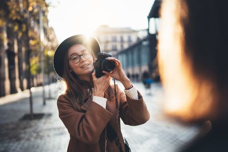 Photographer in glasses with retro camera take photo girlfriend. Tourist smiling girl in hat travels in Barcelona holiday with friend. Sunlight street in europe city. Traveler hipster shoot girlsの写真素材