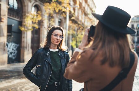 Photographer in glasses with camera take photo model girlfriend. Tourist smiling girl in hat travels in Barcelona holiday with traveler friend. Sunlight flare street in europe city. Photoshoot conceptの写真素材