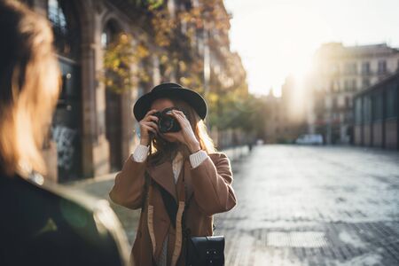Blogger photoshoot concept. Photographer with camera take photo model girlfriend. Tourist smiling girl travels in Barcelona holiday with traveler friend. Sunlight flare street in europe cityの写真素材