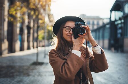 Outdoor smiling lifestyle portrait of pretty young woman having fun in sun city in Europe with camera travel photo of photographer Making pictures in hipster style glasses and hatの写真素材