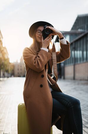 Photographer tourist with suitcase take photo on retro camera. Smiling girl in hat travels in Barcelona. Sunlight flare street in europe city. Traveler hipster shooting architecture, copy space mockupの写真素材
