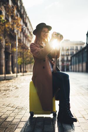 Photographer tourist with suitcase take photo on retro camera. Smiling girl in hat travels in Barcelona. Sunlight flare street in europe city. Traveler hipster shooting architecture, copy space mockupの写真素材