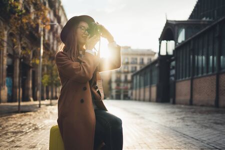 Photographer tourist with suitcase take photo on retro camera. Girl in hat travels in Barcelona holiday. Sunlight flare street in europe city. Traveler hipster shooting architecture, copy space mockupの写真素材