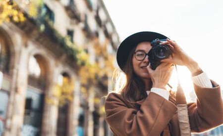 Photographer in glasses with retro photo camera. Tourist portrait. Smiling girl in hat travels in Barcelona holiday. Sunlight flare street in europe city. Traveler hipster shoot architecture, copy space mockupの写真素材