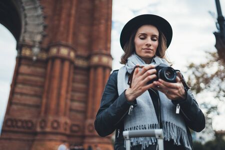 Photographer tourist with retro photo camera. Girl in hat travels in Triumphal arch Barcelona. Holiday concept street in europe city. Traveler hipster shooting architecture, copy space mockupの写真素材