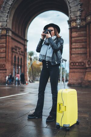 Photographer tourist with retro photo camera. Girl in hat travels in Triumphal arch Barcelona. Holiday concept in europe city street . Traveler hipster shooting architecture, copy space mockupの写真素材