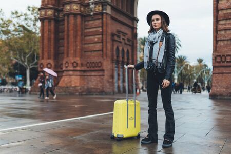 Photographer tourist with retro photo camera. Girl in hat with suitcase travels in Triumphal arch Barcelona. Holiday concept in europe street. Traveler hipster shoot architecture cityの写真素材