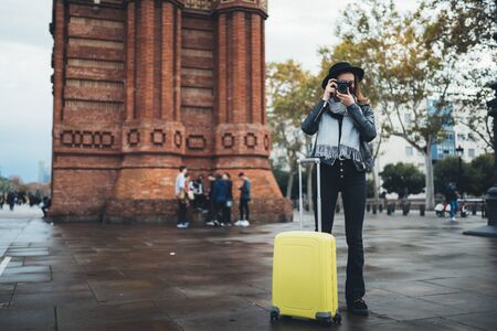 Photographer tourist with retro photo camera. Girl in hat with suitcase travels in Triumphal arch Barcelona. Holiday concept in europe street. Traveler hipster shoot architecture cityの写真素材