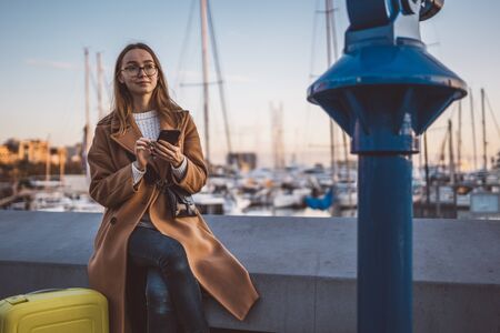 Traveler woman with suitcase holding mobile phone in evening port Barcelona. Girl tourist in glasses using smartphone technology online gadget cellphone. Digital internet lifestyle mockupの写真素材