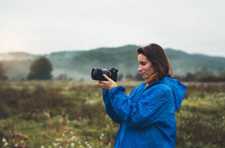 photographer tourist girl in blue raincoat hold in female hands photo camera take photography foggy mountain, traveler shooting autumn nature, video click on camera technology, journey landscape vacation concept free spaceの写真素材
