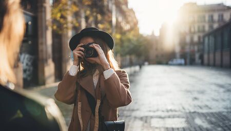 Sunlight flare street in europe city. Photographer in glasses with retro camera take photo model girlfriend. Tourist smiling girl in hat travels in Barcelona holiday. Photoshoot conceptの写真素材