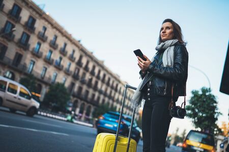 Traveler woman with suitcase calling mobile phone waiting yellow taxi in evening street europe city Barcelona. Girl tourist using smartphone wifi internet online gadget cellphoneの写真素材