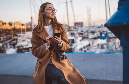 Traveler woman calling mobile phone in evening sea port Barcelona. Girl tourist in glasses using smartphone technology online gadget cellphone. Digital communicationの写真素材