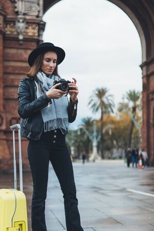Photographer tourist with retro photo camera. Young girl in hat travels in Triumphal arch Barcelona. Holiday concept street in europe city. Traveler hipster shooting architecture, copy space mockupの写真素材