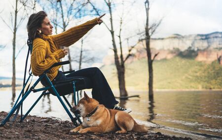 tourist traveler girl relax drink tea together dog on background mountain, puppy pet woman shows hand on lake shore nature trip, friendship conceptの写真素材