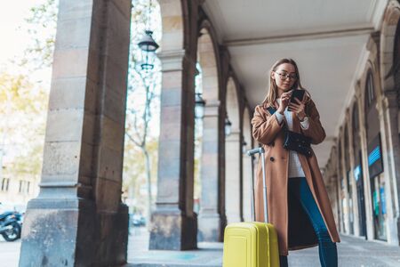 Tourist with suitcase holding smart mobile phone. Girl travels in city Barcelona. Holiday concept in Spain street. Traveler hipster in glasses using cellphone internet technology in europe trip. Copy space mockupの写真素材