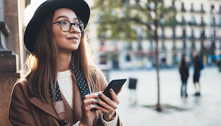 Traveler woman in hat hold in hands mobile phone. Smartphone technology internet online. Girl tourist in glasses using gadget cellphone in sun city. Digital lifestyle communicationの写真素材
