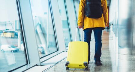 traveler with yellow suitcase backpack at airport on background window blue sky, passenger waiting flight in departure lounge area, airport lobby terminal, vacation trip conceptの写真素材