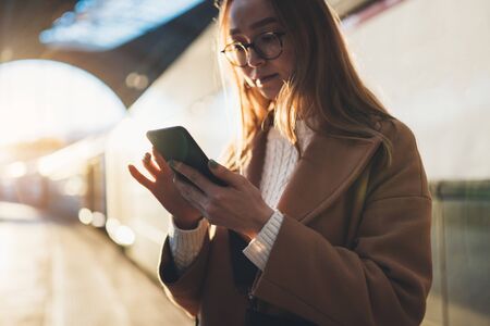 Woman holding in hands mobile phone on platform station. Technology smartphone online connect. Girl travel waiting train using gadget cellphone. Digital wifi internet lifestyle mockupの写真素材